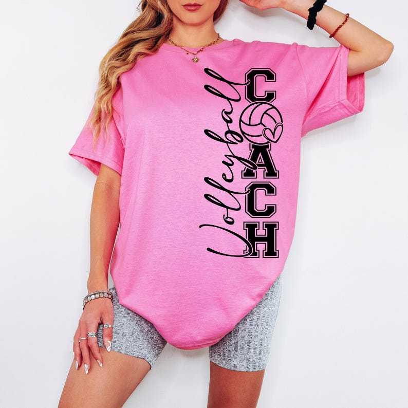 K&ouml;nnte beinhalten: Pinkes T-Shirt mit einem schwarzen Grafikdesign, auf dem "Volleyball Coach" mit einem Volleyball und einem Herz steht.