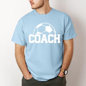 May include: Light blue t-shirt with a white soccer ball graphic and the word "COACH" printed in white.