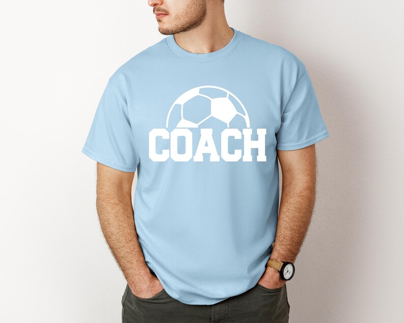 May include: Light blue t-shirt with a white soccer ball graphic and the word "COACH" printed in white.
