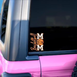 May include: A pink vehicle with a black window displaying a sticker that says "Basketball Mom." The sticker features the word "Basketball" in a script font, a basketball graphic, and the word "MOM" in block letters.