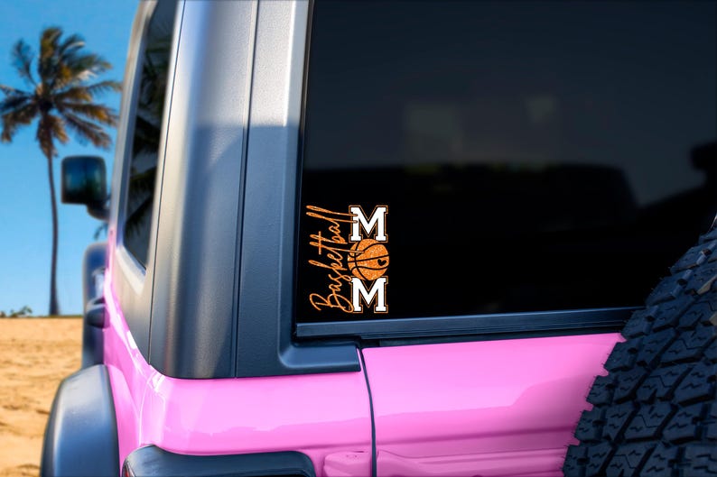 May include: A pink vehicle with a black window displaying a sticker that says "Basketball Mom." The sticker features the word "Basketball" in a script font, a basketball graphic, and the word "MOM" in block letters.