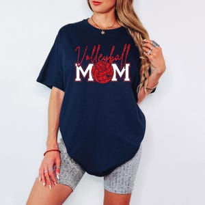 Peut inclure: T-shirt bleu marine avec l'inscription "Volleyball Mom" en paillettes rouges et un graphisme de ballon de volley-ball rouge &agrave; paillettes. Le mot "MOM" est en lettres blanches. Le t-shirt est de style d&eacute;contract&eacute;.