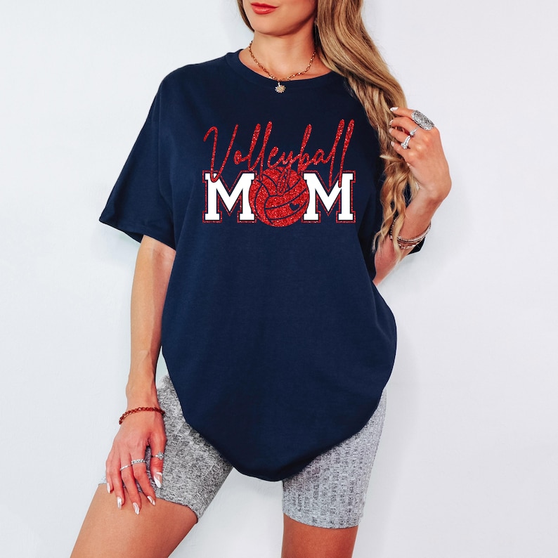 Peut inclure: T-shirt bleu marine avec l'inscription "Volleyball Mom" en paillettes rouges et un graphisme de ballon de volley-ball rouge &agrave; paillettes. Le mot "MOM" est en lettres blanches. Le t-shirt est de style d&eacute;contract&eacute;.