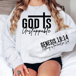 May include: Light gray sweatshirt with the text "GOD IS Unstoppable" and a cross graphic. The sleeve features the text "GENESIS 18:14 Nothing is too hard for God" in black. The sweatshirt is a casual, comfortable style.