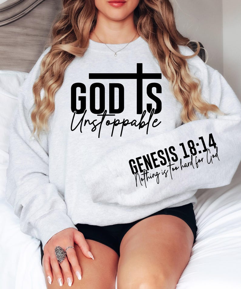 May include: Light gray sweatshirt with the text "GOD IS Unstoppable" and a cross graphic. The sleeve features the text "GENESIS 18:14 Nothing is too hard for God" in black. The sweatshirt is a casual, comfortable style.