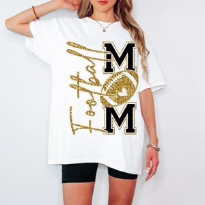 May include: White t-shirt with a gold glitter design. The design features the words "Football Mom" in a script font, with the word "MOM" in black block letters. A gold football is in the center of the design.