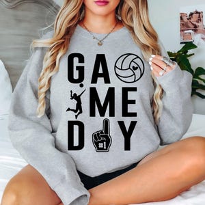 May include: A grey sweatshirt featuring the text "GAME DAY" in black. The design incorporates a volleyball, a player silhouette, and a foam finger. The sweatshirt is a casual garment.