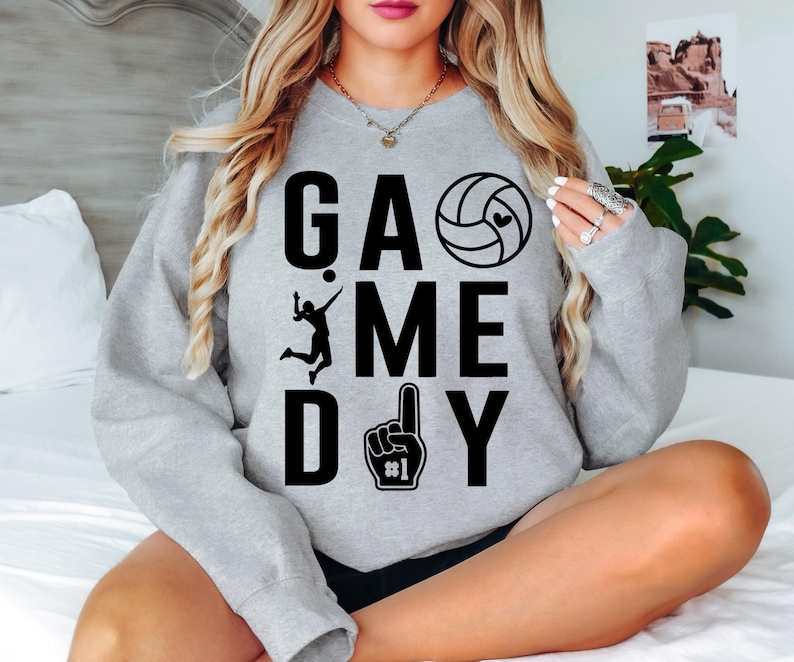 May include: A grey sweatshirt featuring the text "GAME DAY" in black. The design incorporates a volleyball, a player silhouette, and a foam finger. The sweatshirt is a casual garment.