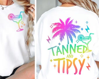 May include: White sweatshirt with a colorful margarita graphic on the front and a palm tree design on the back. The back of the sweatshirt has the words "Tanned and Tipsy" in a rainbow gradient.