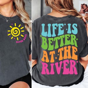 May include: A dark gray t-shirt with a yellow sun graphic on the front and the text "River Days" in pink and white. The back of the shirt has the text "Life is Better at the River" in blue, green, orange, and pink.