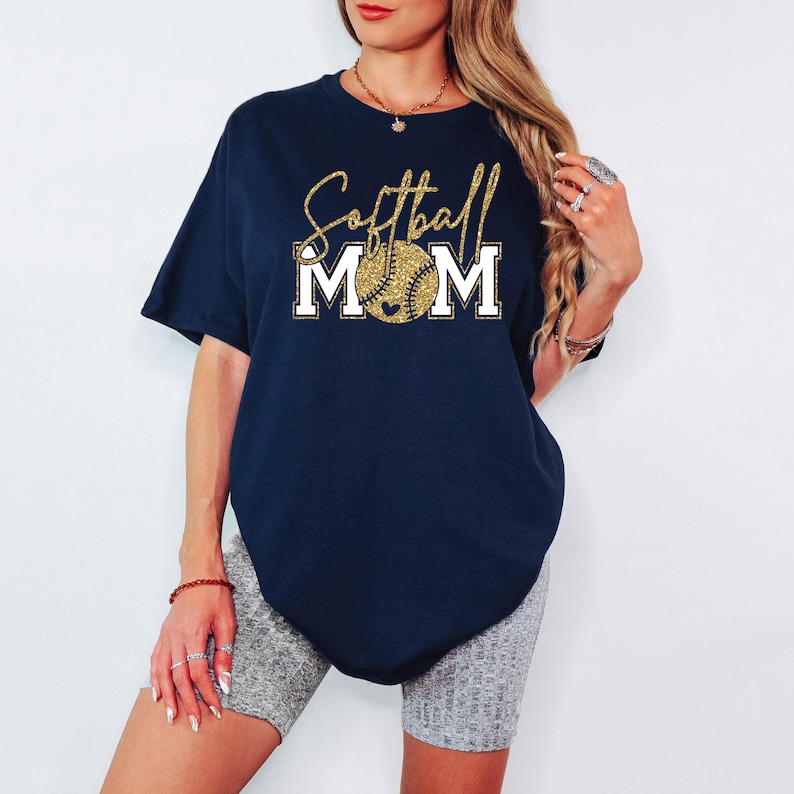 May include: Navy blue t-shirt with the words "Softball Mom" in gold glitter script and a softball graphic. The word "MOM" is in white block letters. A casual tee for softball fans.