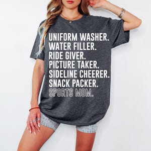 May include: A dark gray t-shirt with white text that reads "UNIFORM WASHER. WATER FILLER. RIDE GIVER. PICTURE TAKER. SIDELINE CHEERER. SNACK PACKER. SPORTS MOM."