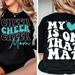 May include: Black t-shirt with a teal glitter design. The front of the shirt says "Cheer Cheer Cheer Mom" and the back says "My heart is on that mat" with a teal glitter heart.