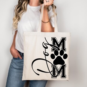 Puede incluir: Una bolsa de lona blanca con una huella de pata negra y el texto "Dog Mom" en una fuente de estilo universitario.