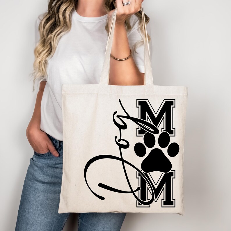 Puede incluir: Una bolsa de lona blanca con una huella de pata negra y el texto "Dog Mom" en una fuente de estilo universitario.