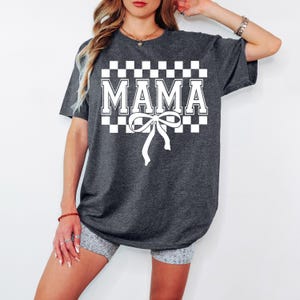 May include: A dark gray t-shirt with a white checkered pattern and the word "MAMA" in white letters. A white bow is centered below the text.