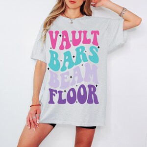May include: A grey t-shirt with the words "VAULT BARS BEAM FLOOR" printed in pink, blue, purple, and white.