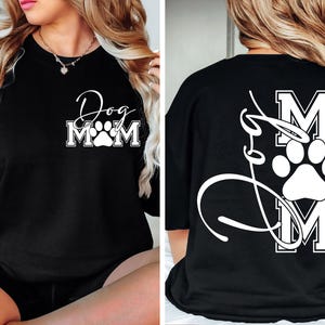Puede incluir: Camiseta negra con una huella de pata blanca y el texto "Dog Mom" en la parte delantera. La parte posterior de la camiseta tiene una huella de pata blanca y el texto "Dog Mom" en una fuente de escritura.