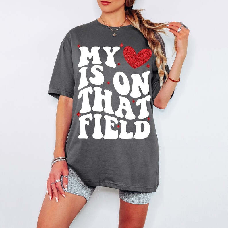 Pu&ograve; includere: Una maglietta oversize di colore grigio scuro con il testo "MY IS ON THAT FIELD" in bianco e un cuore rosso glitterato.