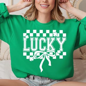 Pu&ograve; includere: Felpa verde con un motivo a scacchi bianchi, un fiocco bianco e la parola "LUCKY" in lettere bianche.