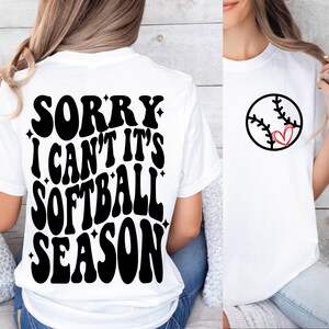 May include: White t-shirt with black text that reads "Sorry I can't it's softball season" on the back. The front of the shirt has a black and white softball with a red heart in the middle.