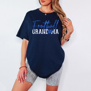Pu&ograve; includere: T-shirt blu navy con la scritta "Football Grandma" in glitter blu. La parola "Grandma" &egrave; in lettere bianche. Una grafica di football sostituisce la lettera "O".