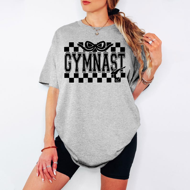 May include: Gray oversized t-shirt with a black and white checkered design and the text "GYMNAST" in black. The design also includes a black ribbon and a gymnast silhouette.