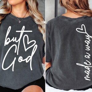 May include: A charcoal gray t-shirt with white text on the front that reads "but God" with a heart symbol. The back of the shirt has white text that reads "made a way".
