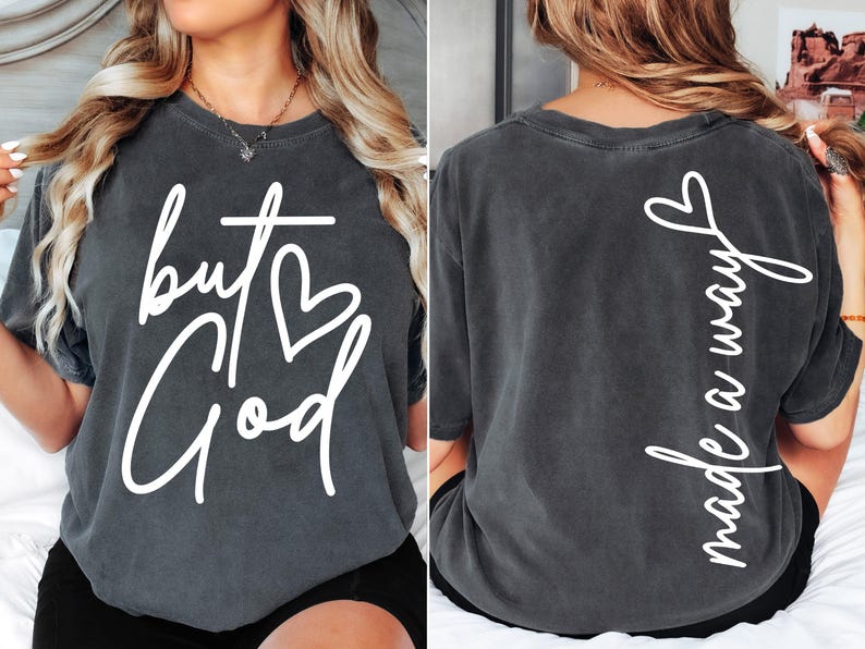 May include: A charcoal gray t-shirt with white text on the front that reads "but God" with a heart symbol. The back of the shirt has white text that reads "made a way".