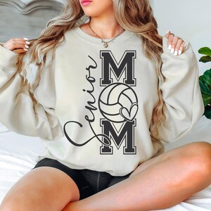 May include: A cream-colored sweatshirt with the words "Senior Mom" and a volleyball graphic printed on the front.