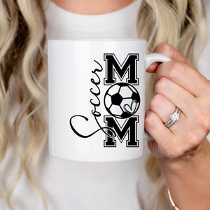 May include: White ceramic mug with a black and white design that says "Soccer Mom" with a soccer ball and heart graphic.