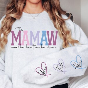 May include: Gray sweatshirt with the text "This Mamaw wears her heart on her sleeve" in pink, purple, and blue. Three heart outlines with the text "Your", "Text", and "Here" are on the sleeves.