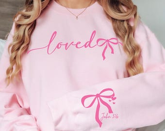 May include: Pink sweatshirt with the word "loved" and a pink bow on the front. The left sleeve has a pink bow with hearts and the text "John 3:16" printed on it.