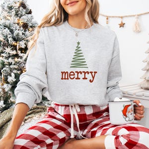 May include: A light gray sweatshirt with a green Christmas tree design and the word "merry" in red. The person is wearing red and white plaid pajama pants and holding a white mug with a Santa Claus graphic.