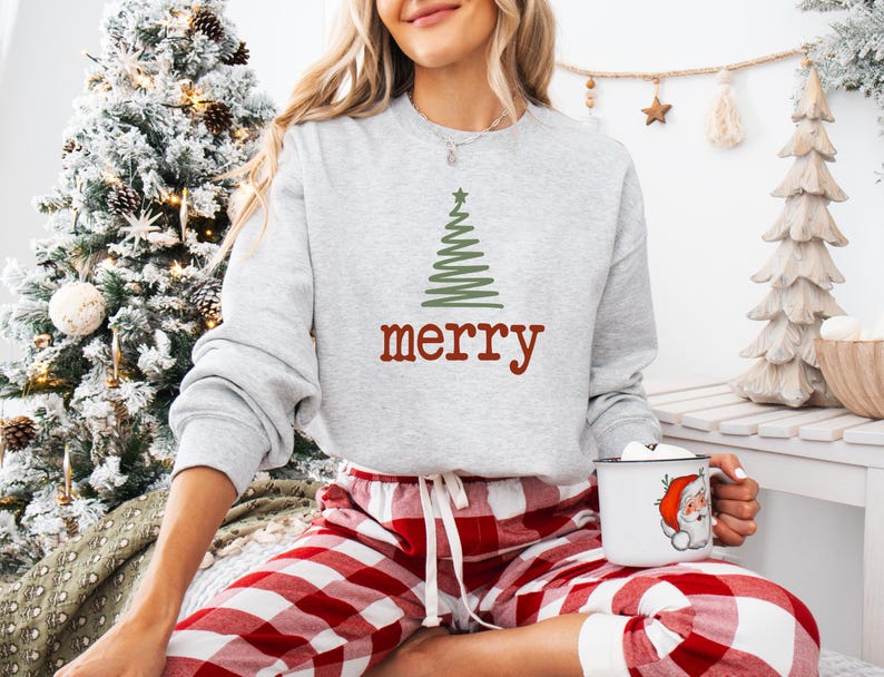 May include: A light gray sweatshirt with a green Christmas tree design and the word "merry" in red. The person is wearing red and white plaid pajama pants and holding a white mug with a Santa Claus graphic.