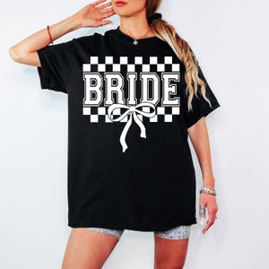 Puede incluir: Camiseta negra con la palabra "BRIDE" en blanco, sobre un fondo a cuadros. Una cinta blanca est&aacute; centrada debajo de la palabra. La camiseta es de estilo casual.