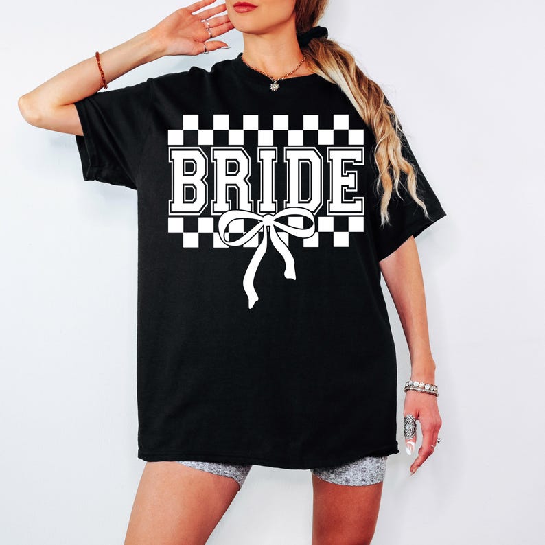 Puede incluir: Camiseta negra con la palabra "BRIDE" en blanco, sobre un fondo a cuadros. Una cinta blanca est&aacute; centrada debajo de la palabra. La camiseta es de estilo casual.