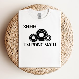 以下が含まれることがあります： 黒い文字で「SHHH... I'M DOING MATH」と書かれた白いTシャツと、10、25、45の数字が書かれた3枚のウェイトプレートのグラフィック。