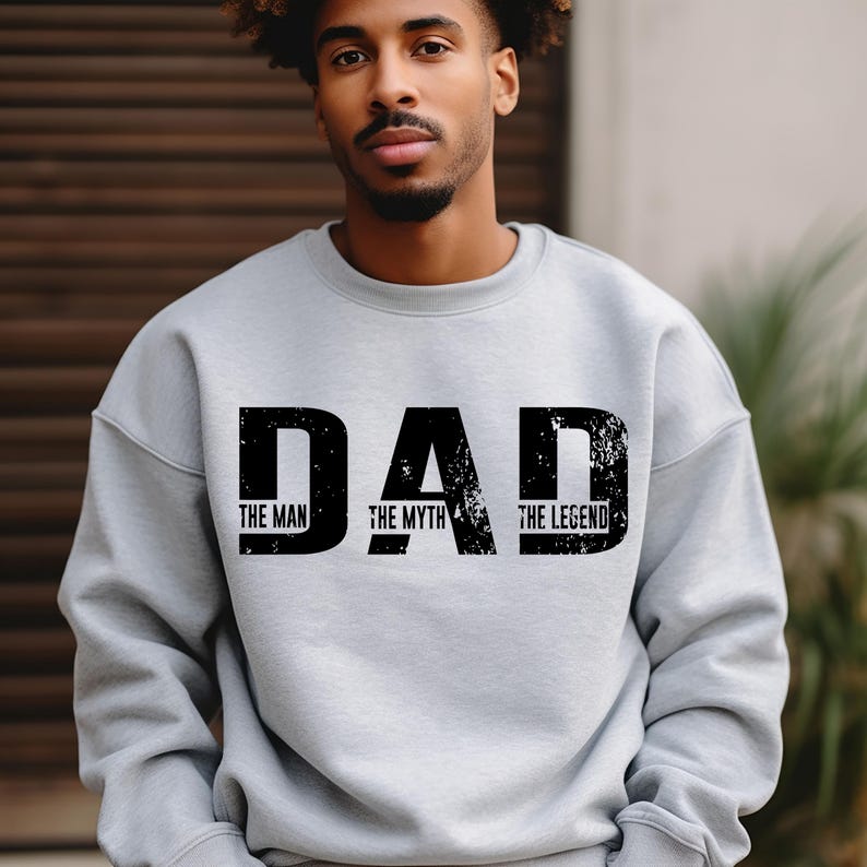 K&ouml;nnte beinhalten: Ein hellgraues Sweatshirt mit Rundhalsausschnitt und dem schwarzen, abgenutzten Schriftzug DAD in gro&szlig;en Gro&szlig;buchstaben. Unter jedem Buchstaben sind die W&ouml;rter THE MAN, THE MYTH und THE LEGEND in kleiner, schwarzer Schrift aufgedruckt.