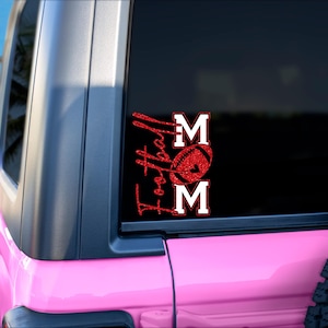 May include: A red glitter vinyl decal on a black car window. The decal reads "Football Mom" with a football graphic. The car is pink and parked on a sandy beach with palm trees in the background.