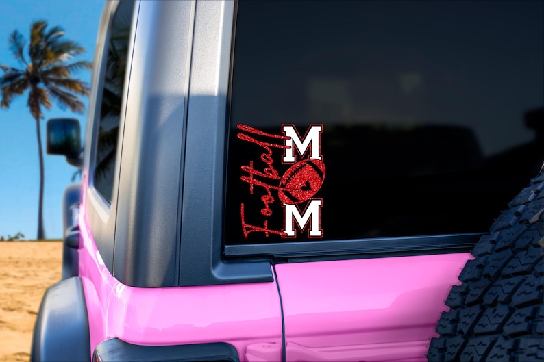 May include: A red glitter vinyl decal on a black car window. The decal reads "Football Mom" with a football graphic. The car is pink and parked on a sandy beach with palm trees in the background.