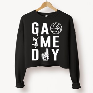 May include: Black cropped sweatshirt with the words "GAME DAY" in white block letters. A volleyball, a volleyball player, and a number one hand gesture are also in white. The sweatshirt is hanging on a wooden hanger.