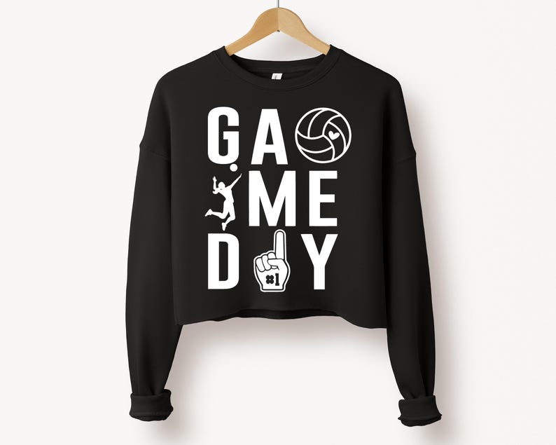 May include: Black cropped sweatshirt with the words "GAME DAY" in white block letters. A volleyball, a volleyball player, and a number one hand gesture are also in white. The sweatshirt is hanging on a wooden hanger.