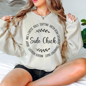 May include: A beige sweatshirt with the text "Side Chick" in a circle surrounded by a list of side dishes, including mashed potatoes, gravy mac & cheese, cheese rolls, stuffing, green bean casserole, and cranberry sauce.
