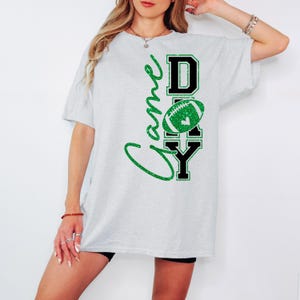 K&ouml;nnte beinhalten: Hellgraues T-Shirt mit den Worten "Game Day" in gr&uuml;nem Glitzerschriftzug und Blockbuchstaben. Ein gr&uuml;ner Football mit einem Herz befindet sich in der Mitte. Das Shirt hat einen l&auml;ssigen, &uuml;bergro&szlig;en Stil.