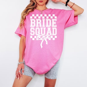 Puede incluir: Una camiseta rosa con las palabras "BRIDE SQUAD" en letras blancas. El texto est&aacute; enmarcado por un patr&oacute;n de cuadros blancos y grises. Una cinta blanca est&aacute; debajo del texto. La camiseta es de corte holgado.