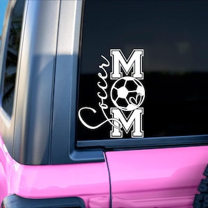 May include: A white vinyl decal with the words "Soccer Mom" and a soccer ball with a heart in the middle. The decal is on the back window of a pink car.