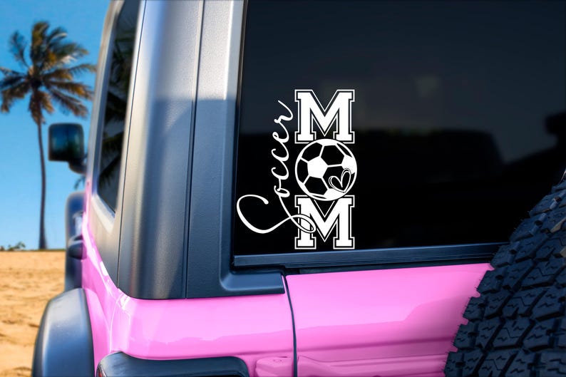 May include: A white vinyl decal with the words "Soccer Mom" and a soccer ball with a heart in the middle. The decal is on the back window of a pink car.