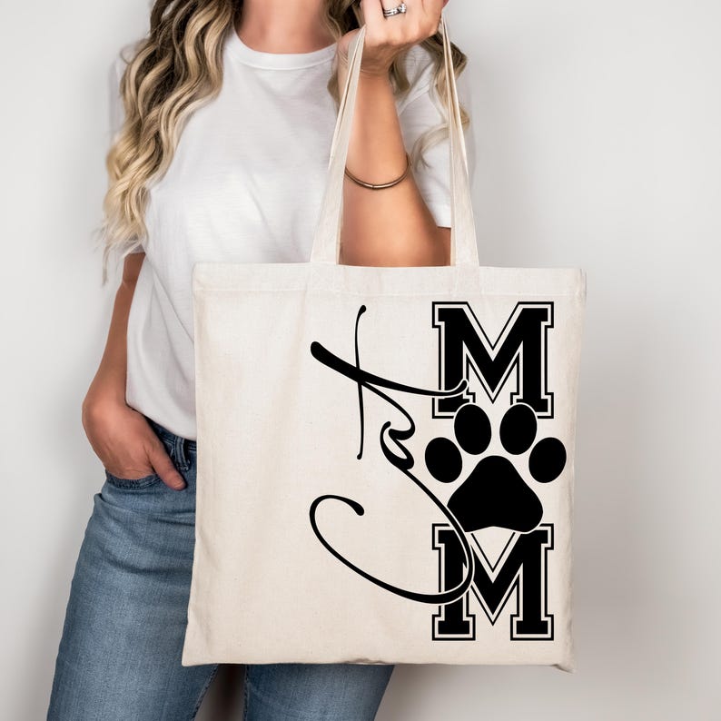 May include: Cream-colored tote bag with black text and a paw print design. The text reads "Dog Mom" in a stylized font. The bag has two handles and is held by a person.