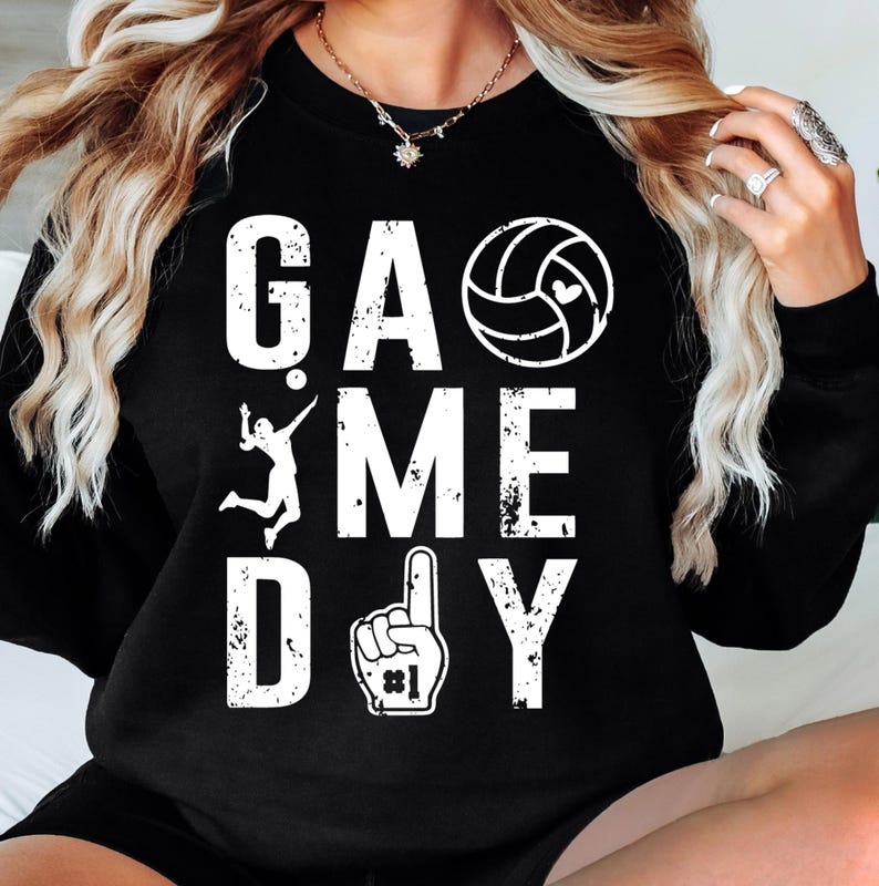 May include: Black sweatshirt featuring the text "GAME DAY" in white, accompanied by a volleyball graphic, a volleyball player silhouette, and a foam finger. The design has a distressed, vintage appearance.